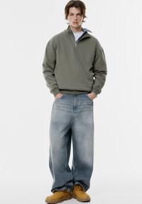 Maglione verde oliva chiaro con zip a un quarto, abbinato a jeans larghi blu chiaro e stivali gialli. Tessuto morbido, vestibilità rilassata e design semplice.