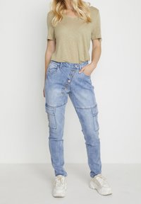 Vaaleansiniset farkkucargo-housut, joissa on useita taskuja, hieman kapeneva leikkaus ja sivunappikiinnitys. Yhdistettynä khaki t-paitaan ja valkoisiin lenkkareihin.