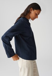 Camicia a maniche lunghe blu navy con colletto alto, polsini con bottoni e vestibilità rilassata, realizzata in tessuto morbido. Abbinata a pantaloni beige chiaro.
