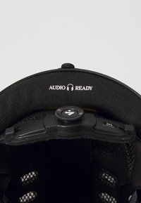 Melns ķivere iekšpuse ar mīkstu audumu, regulējams diapazons un teksts "AUDIO READY". Iekļauj mazas ventilācijas atveres un izmēra marķējumu M/L.