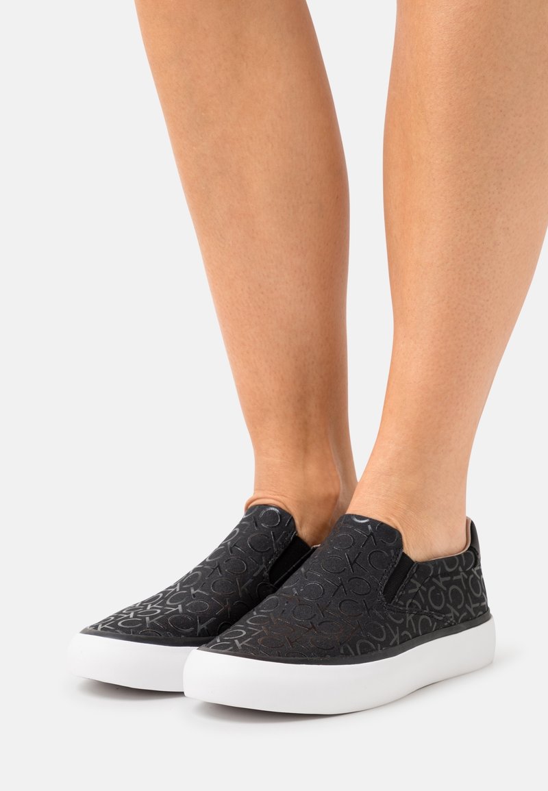 Baskets noires sans lacets avec semelles blanches, présentant un motif subtil « CK » sur l’ensemble, portées pieds nus sur un fond clair uni.