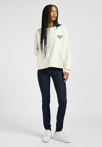 Kremowy sweatshirt z wzorem serca, zestawiony z ciemnymi dżinsami i białymi trampkami. Sweatshirt ma luźny krój i ściągacze przy rękawach.