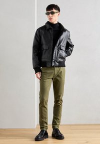 Jachetă din piele neagră cu buzunare aplicate, pantaloni verzi oliva ajustați și pantofi negri. Modelul poartă ochelari de soare verzi și un pulover negru cu gât.
