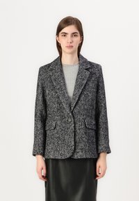 Blazer noir et blanc à motif pied-de-poule avec un seul bouton, poches à rabat et revers crantés ; porté sur un pull en maille côtelée gris.