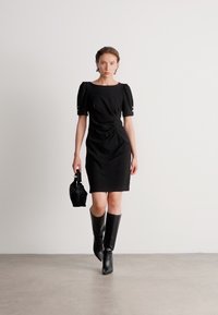 Crna tijesna haljina s kratkim napuhanim rukavima, kvadratnim dekolteom i nabiranjem sa strane, uparena s čizmama do koljena i malom torbicom.