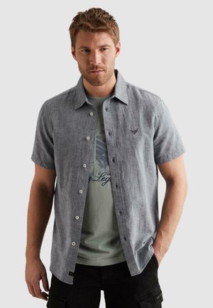 Homme aux cheveux châtain clair portant une chemise gris clair à manches courtes boutonnée sur un T-shirt vert imprimé et un pantalon noir.