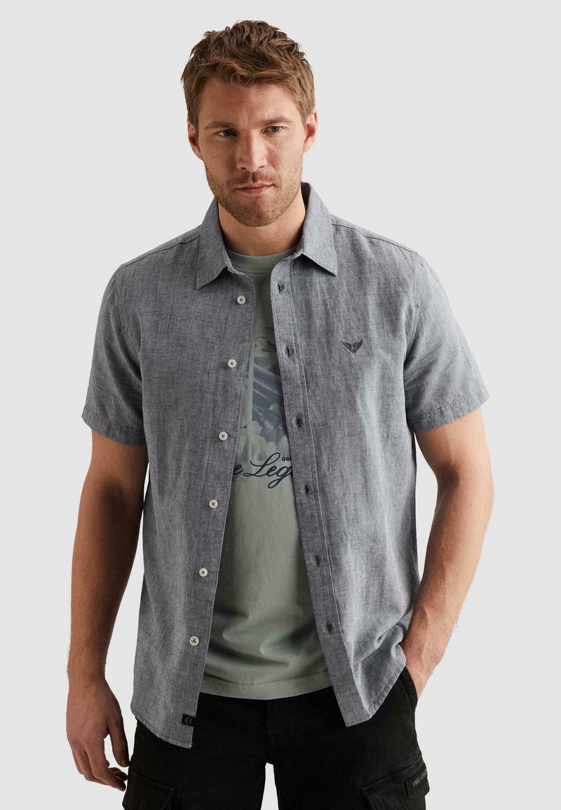 Homme aux cheveux châtain clair portant une chemise gris clair à manches courtes boutonnée sur un T-shirt vert imprimé et un pantalon noir.