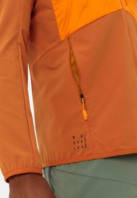 Giacca in tessuto riciclato con una sezione superiore arancione, una tasca con zip e dettagli in rete, e un polsino elasticizzato per il comfort.