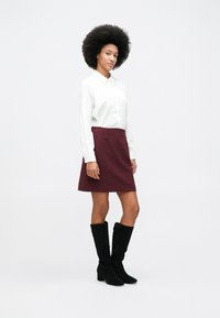 Woman with afro hairstyle wearing a white long-sleeve shirt, burgundy skirt, and black knee-high boots, standing against a plain white background.