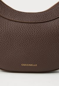Borsa in pelle marrone con una finitura texturizzata, caratterizzata da una forma curva e un logo dorato "COCCINELLE" nella parte inferiore. Accentuata da dettagli cuciti.