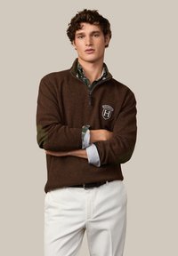 Junger Mann mit lockigem Haar, der einen braunen Pullover mit Viertelreißverschluss und Logo über einem grünen karierten Hemd sowie weiße Hosen trägt, die Arme verschränkt.