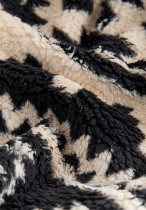 Weiche, strukturierte Decke mit beigem Grund und kontrastierenden schwarzen abstrakten Mustern. Das Material wirkt plüschig mit einem flauschigen Finish.