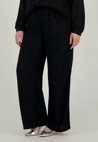 Άνθρωπος που φοράει φαρδιά μαύρα παντελόνια με απαλές λάμψεις και ασημένιες μυτερές γόβες, στέκεται σε έναν απλό ανοιχτό φόντο.