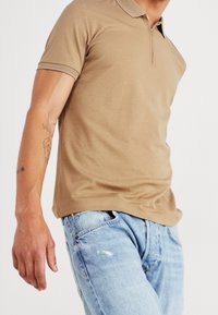 Tan polo skjorta med dragkedja och kontrasterande strukturerade detaljer. Bärs med ljusblå slitna jeans. Slät tyg, figursydd design.