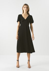 Musta midi-mekko, jossa on V-pääntie, lyhyet frillat-sleet ja virtaava siluetti. Sujuva kangas yksinkertaisella, minimalistisella muotoilulla.