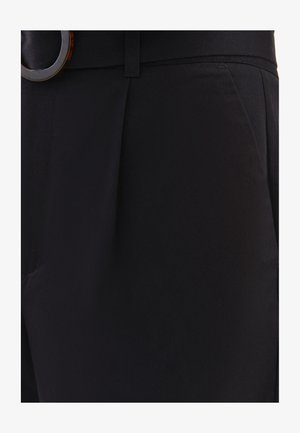 Los pantalones negros ajustados cuentan con una textura suave, una cintura estructurada, pliegues sutiles y un accesorio de hebilla de cinturón en forma circular de carey.