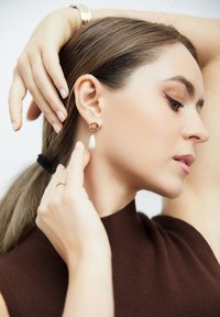 Pendientes de aro dorados con una perla colgante, llevados por una modelo con cabello largo y castaño, que lleva una prenda marrón acanalada y un fondo neutro.