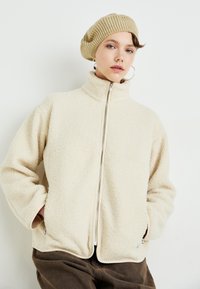Young person wearing beige knit beret, hoop earrings, cream fleece zip-up jacket, and brown pants, standing against plain background.