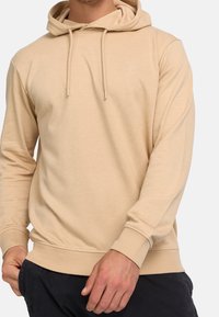 Sweat à capuche beige en tissu doux avec un cordon de serrage, des poignets et un ourlet côtelés ; présente un logo discret sur la poitrine.