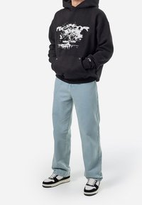 Svart huvtröja med grafiskt tryck, kombinerad med ljusblå jeans och svarta och vita sneakers. Avslappnad, lös passform.