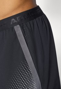 Zwarte sportshorts hebben een gestructureerde elastische tailleband met "ARMOUR" branding, een zijpaneel met mesh en een stippenpatroon, en zijn gemaakt van een zachte stof.