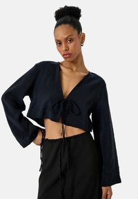 Granatowa, krótka bluzka z długimi rękawami, głębokim dekoltem w kształcie litery V i szczegółem w postaci wiązania z przodu. Teksturowany materiał, zestawiona z czarnymi spodniami z troczkami.