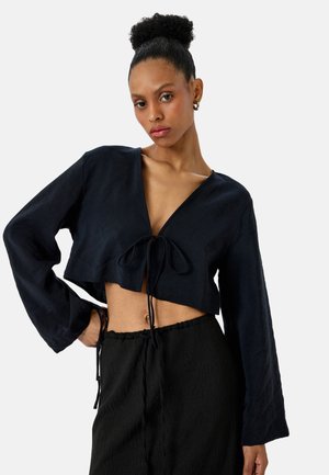 Granatowa, krótka bluzka z długimi rękawami, głębokim dekoltem w kształcie litery V i szczegółem w postaci wiązania z przodu. Teksturowany materiał, zestawiona z czarnymi spodniami z troczkami.