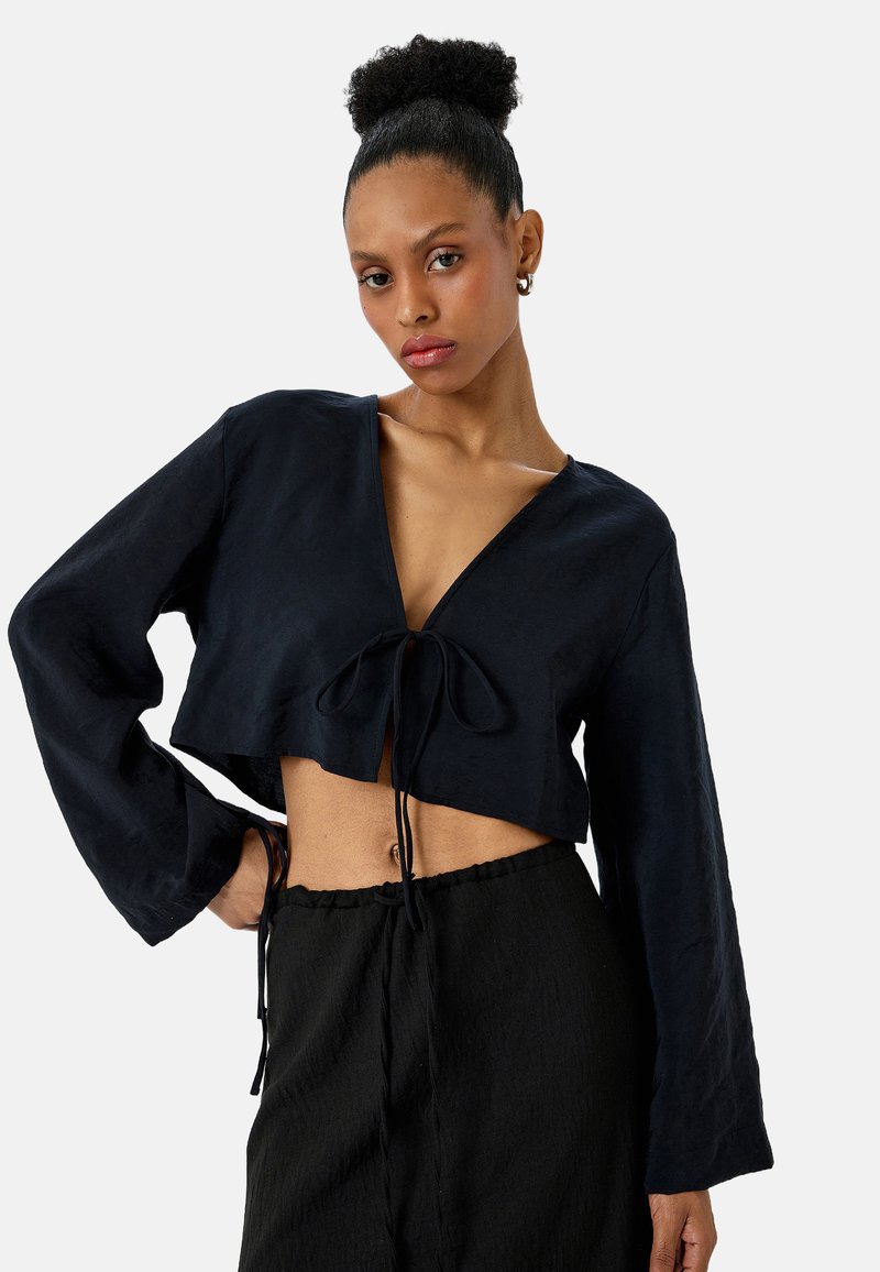 Granatowa, krótka bluzka z długimi rękawami, głębokim dekoltem w kształcie litery V i szczegółem w postaci wiązania z przodu. Teksturowany materiał, zestawiona z czarnymi spodniami z troczkami.