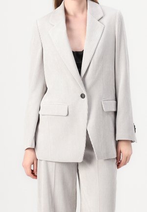 Vrouw die een lichtgrijze corduroy blazer draagt met een eenknopsluiting en bijpassende wijde broekspijpen, gecombineerd met een zwart topje.