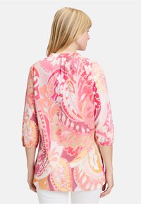 Blouse colorée à motifs présentant des designs paisley roses, oranges et jaunes, avec des manches 3/4 et un ourlet arrondi à l'arrière. Fabriquée en tissu léger.