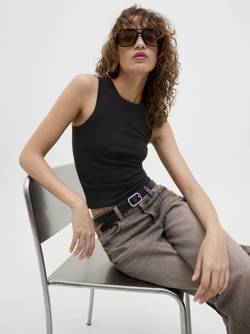 Top senza maniche nero, design aderente. Jeans grigio-marroni con una cintura nera. Modella con capelli ricci, che indossa occhiali da sole neri oversize, seduta su una sedia di metallo.