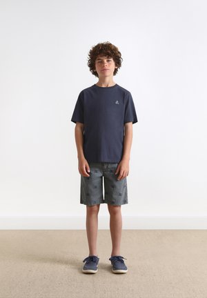 Marineblaue Baumwoll-T-Shirt mit kurzen Ärmeln und kleinem Logo auf der Brust, kombiniert mit grauen Shorts mit einem dezenten Blattmuster und blauen Schuhen.