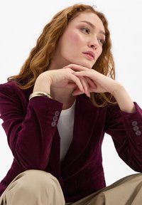 Roodharige vrouw in een bordeauxblauwe blazer en beige broek steunt haar kin op haar verstrengelde handen en kijkt nadenkend naar rechts op een witte achtergrond.