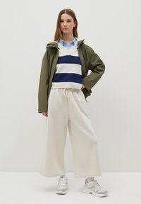 Giacca impermeabile verde oliva, maglione a righe blu e bianche, pantaloni larghi color crema e sneaker bianche chunky con accenti sottili.