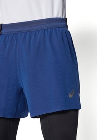 Blauwe sportshorts met een textuurpatroon, voorzien van een zwarte tailleband en een subtiele Asics-logo in goud. Draagt over zwarte compressietights.