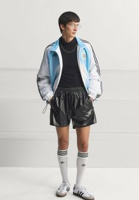 Άνδρας που φοράει αθλητικό μπουφάν Adidas σε ανοιχτό μπλε και λευκό, μαύρο πουλόβερ με ψηλό γιακά, γυαλιστερό μαύρο σορτς, λευκές κάλτσες Adidas μέχρι το γόνατο και λευκά αθλητικά παπούτσια.