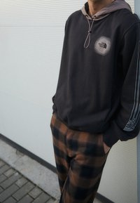 Sudadera con capucha negra y forro beige, con un logo en el pecho y rayas blancas. Combinada con pantalones de cuadros marrones.