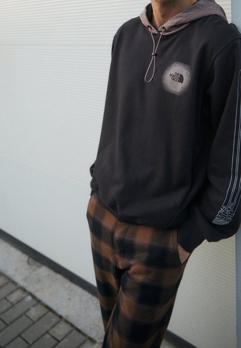 Sudadera con capucha negra y forro beige, con un logo en el pecho y rayas blancas. Combinada con pantalones de cuadros marrones.