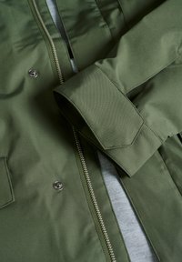 Olivegrüne Jacke mit glatter Textur, Reißverschluss und Druckknöpfen. Verfügt über einen umgeschlagenen Ärmel und ein gefüttertes Innenfutter. Einfaches und funktionales Design.