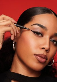 Donna con pelle liscia e orecchini a cerchio argentati applica eyeliner nero a forma di ala sulla palpebra contro uno sfondo rosso.