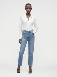 Høj kvinde iført en hvid langærmet knapbluse, lyseblå højtaljede jeans og sorte åbne sandaler stående mod en ensfarvet baggrund.