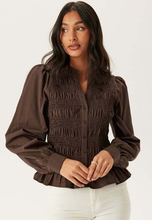 Kvinde med langt mørkt hår iført en brun rynket langærmet bluse og hvide bukser, hænderne foldet ved taljen, kigger mod højre.