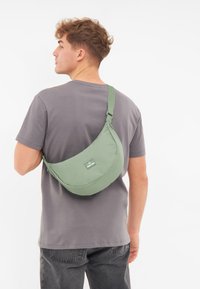 Borsa a tracolla in morbido tessuto verde, forma arrotondata, tracolla regolabile e un piccolo etichetta verde. Indossata sulla spalla, abbinata a una maglietta grigia.