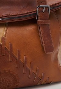 Brun lædertaske med en struktureret overflade, dekorative syninger og prægede cirkulære mønstre. Inkluderer en sølvfarvet lynlås og stropdetalje.