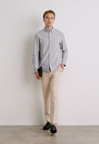 Ανοιχτό γκρι πουκάμισο με κουμπιά, μπεζ παντελόνι, μαύρα παπούτσια, κρατώντας μια ταμπλέτα. Απλός σχεδιασμός, λεία υφή και εφαρμογή κομμένη με ραφή.