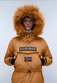 Person iført en brun Napapijri parka med pelsforet hætte trukket tæt, hænderne i lommerne, norsk flagmærke på lommen foran.