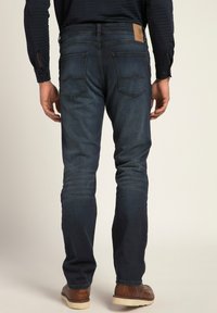 Jeans de mezclilla azul oscuro con un corte clásico, detalles de costura visibles y dos bolsillos traseros. Llevados con zapatos marrones; se muestra la vista trasera.