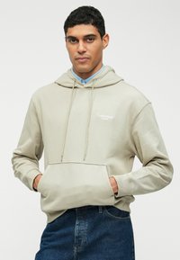 Ανοιχτό γκρι φούτερ με μπροστινή τσέπη, κουκούλα με κορδόνι και λογότυπο Calvin Klein. Φορεμένο πάνω από ένα πουκάμισο με γιακά και συνδυασμένο με μπλε τζιν.