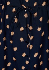 Tissu bleu marine avec des pois beiges irréguliers, présentant un cordon noué et une rangée verticale de boutons noirs au centre.