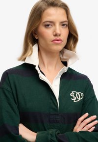 Jeune femme les bras croisés, portant un maillot de rugby rayé vert et noir avec un col blanc relevé et un logo brodé.